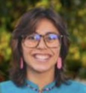 Headshot of Analyse Christine Ramirez smiling in glasses, pink earrings, and blue top with blurred foliage background