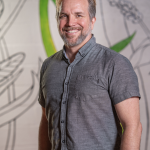 Picture of Brian Lamere smiling standing in front of an artistic back wall with large perhaps unfinished illustrations of grass and plants and what looks to be a dragon fly