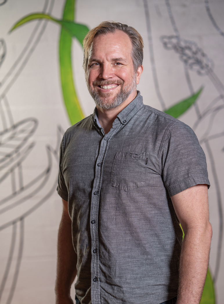 Picture of Brian Lamere smiling standing in front of an artistic back wall with large perhaps unfinished illustrations of grass and plants and what looks to be a dragon fly