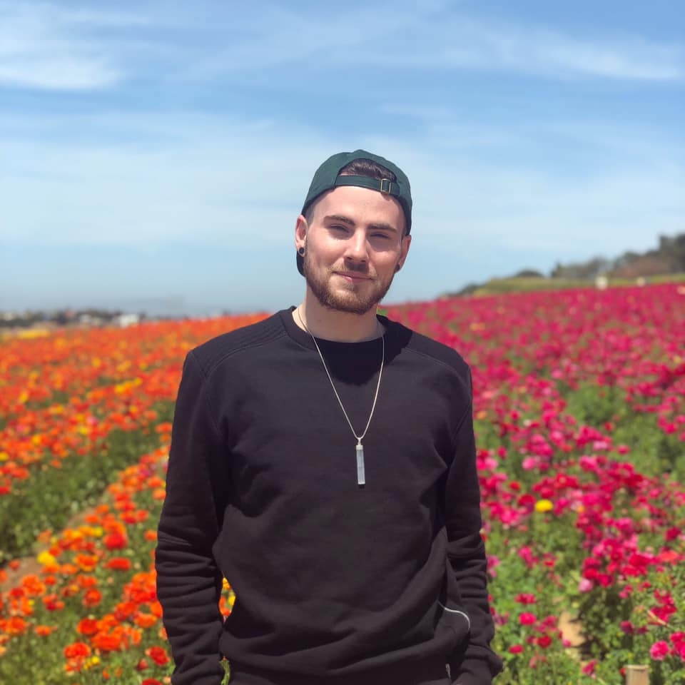 Picture of Casey Kennedy smiling in dark long sleeved top with long thin necklace with pendant and backward cap in front of fields of flowers