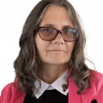 Headshot of Coleen Cusack with fuscia cape, long sleeve black sweater with white collared formal shirt peaking out of the sweater and a gender equality pin wearing glasses on a white background