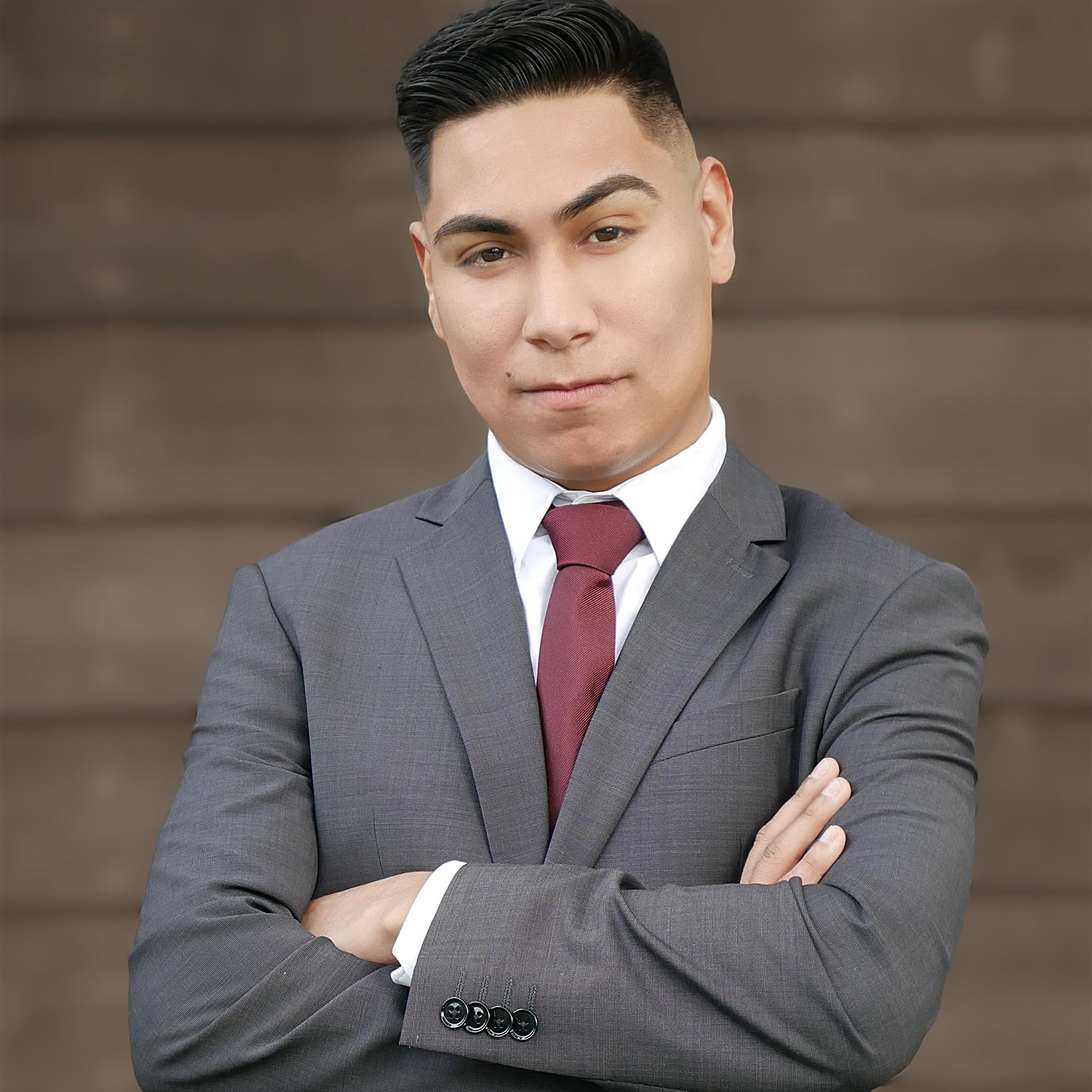 Picture of Danny Avitia with arms folded in a grey business jacket and maroon tie with white collared shirt against a blurred building wall
