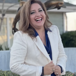 Medium body shot of Delfina Gonzalez smiling in a off white jacket with dark blue top in front of a founatin with bronze dolphins and greenery