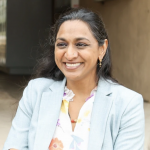 Picture of Dr Darshana Patel in front of a public school with a beaming smile, wearing a light powder blue collared jacket and floral v-neck blouse