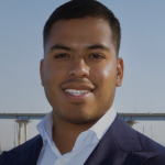 Picture of Japhet Perez Estrada smiling in a navy blue business jacket and open white collared shirt against the Coronado Bridge skyline