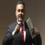 Picture of José Sarmiento in a business suit with red tie and white collared shirt with microphone in hand and papers in the other against a warm grey brownish background