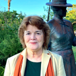 Picture of Kate Callen in a garden with a bronze statue of an early 20th century woman in a wide brimmed hat in the background