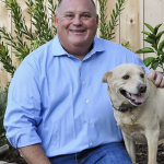 Picture of Kevin Juza smiling in a blue rolled up sleeves collared shirt and jeans, sitting down with a beige medium-sized dog with a plant and wooden fence in the background