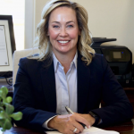 Picture of Kristie Bruce-Lane smiling at a desk with pen in hand wearing a dark navy blue suit jacket and light blue pin-striped collared shirt