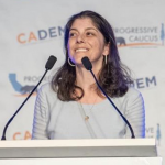 Picture of Lauren Bier smiling behind a podium with two microphones against a backdrop that says "CADEM Progressive Caucus" in a pattern