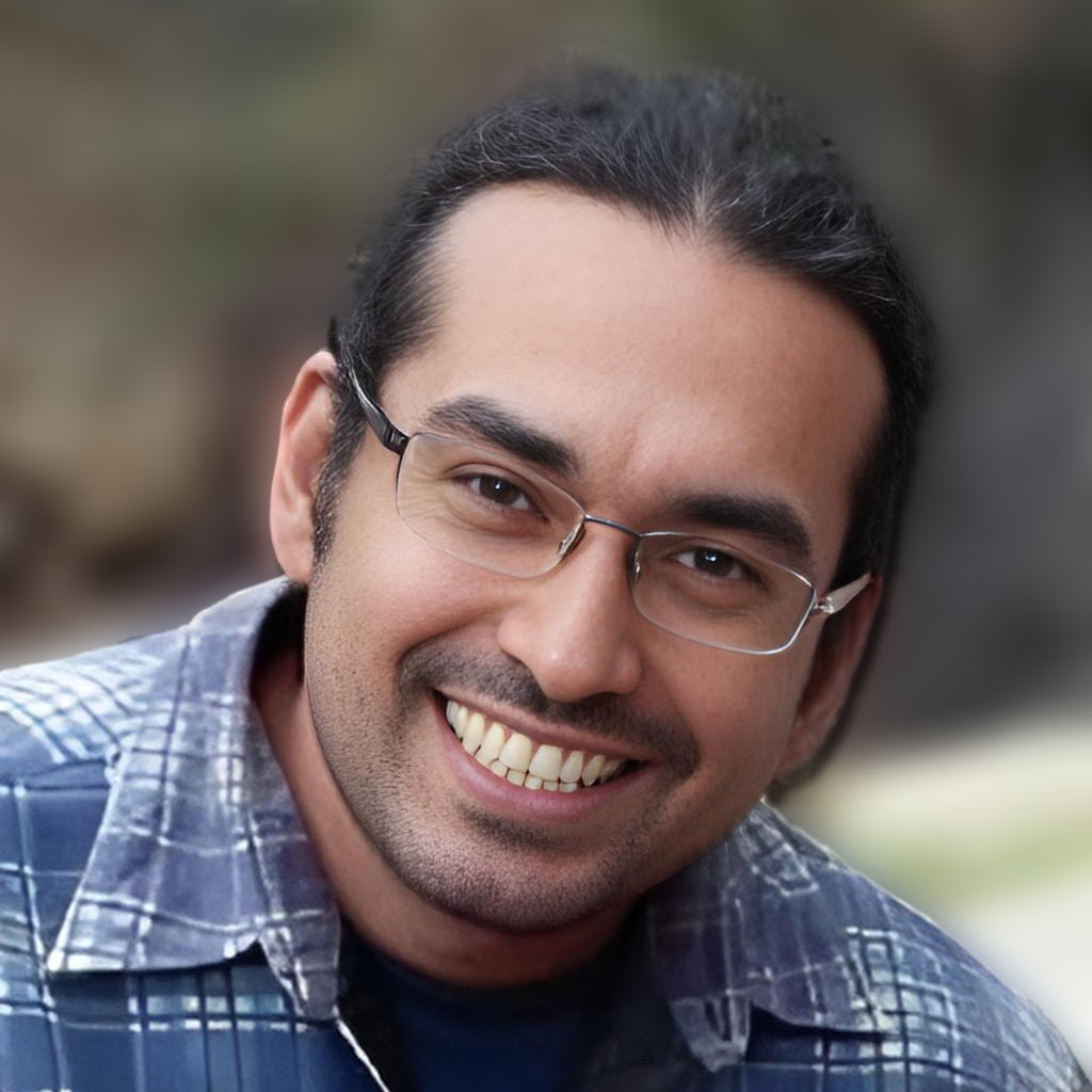Headshot of Manuel Gomez smiling, wearing glasses and a plaid collared shirt
