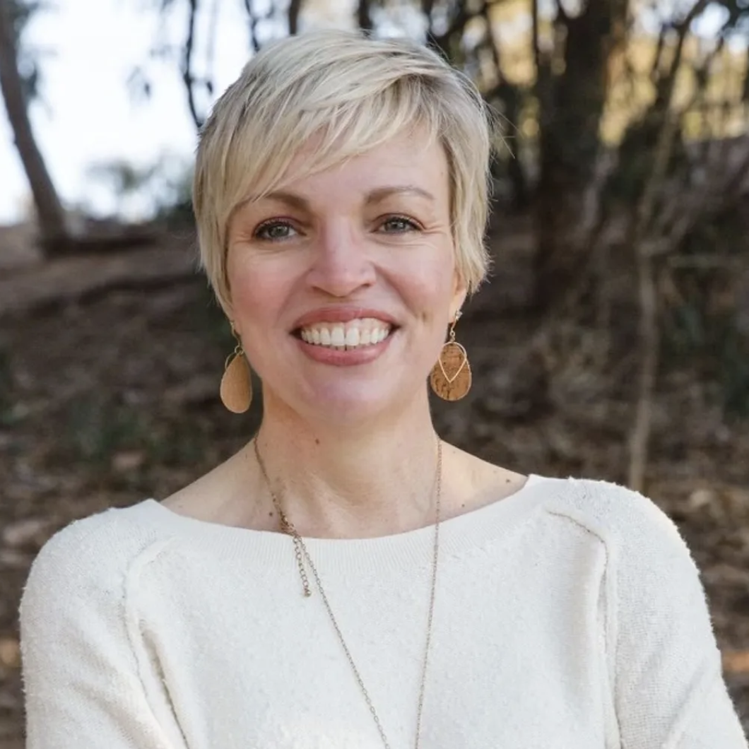 Picture of Nikki Faddick smiling with brown round earrings, cream top, and thin long necklace against a blurrede wooded area