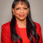 Picture of Tylisa Suseberry in red formal suit on warm grey background, smiling