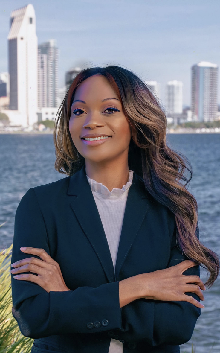 Medium body shot of Geneviéve Jones-Wright with arms folded, smiling with formal navy blue jacket and white blouse with the bay and City of San Diego in the background