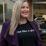 Medium body shot of Alysson Snow outside a building wearing a purple jacket with black shirt beneath with white text saying "run like a girl" smiling and looking to the right