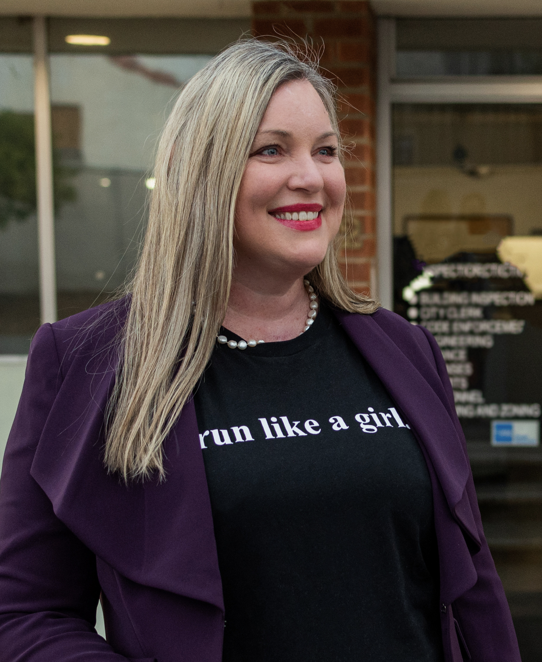 Medium body shot of Alysson Snow outside a building wearing a purple jacket with black shirt beneath with white text saying "run like a girl" smiling and looking to the right