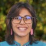 Headshot of Analyse Christine Ramirez smiling in glasses, pink earrings, and blue top with blurred foliage background