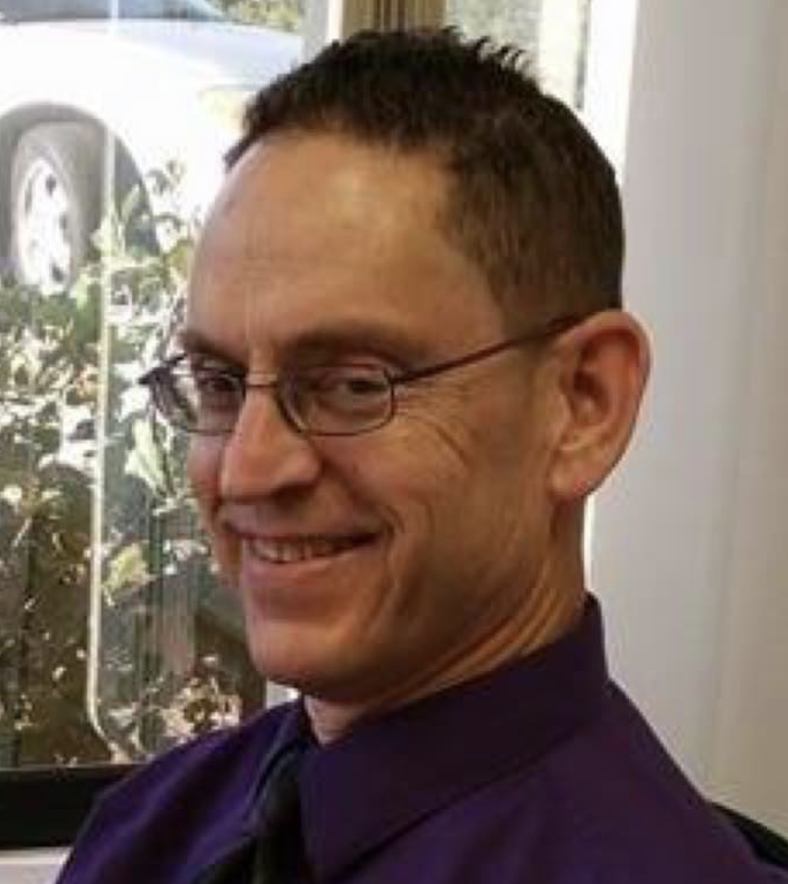 Picture of Brian Polejes smiling wearing glasses and purple shirt and tie in front of a window