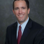 Chris Lawson smiling wearing a black business jacket, red tie, and light blue collared shirt against a grey mottled background