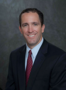 Chris Lawson smiling wearing a black business jacket, red tie, and light blue collared shirt against a grey mottled background