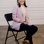 Picture of Destiny Preston smiling and sitting on a black foldable chair with pink collared jacket, white top, and black pants with chunky brown sandals on a polished stone floor and white wall in the background