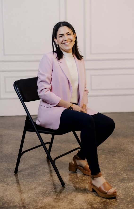 Picture of Destiny Preston smiling and sitting on a black foldable chair with pink collared jacket, white top, and black pants with chunky brown sandals on a polished stone floor and white wall in the background