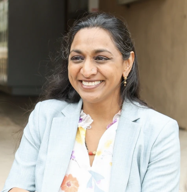 Picture of Dr Darshana Patel in front of a public school with a beaming smile, wearing a light powder blue collared jacket and floral v-neck blouse