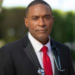 Dr Eugene Allen in a black business jacket, red tie, white collared shirt with a stethoscope against a blurred greenery background
