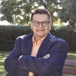 Eric Gonzales smiling with crossed arms in a dark blue business jacket and salmon collared shirt and wearing glasses