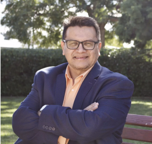 Eric Gonzales smiling with crossed arms in a dark blue business jacket and salmon collared shirt and wearing glasses