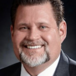 Erik Peterson smiling in a black business jacket wearing a red tie with thin white stripes and a white collared shirt against a dark backdrop