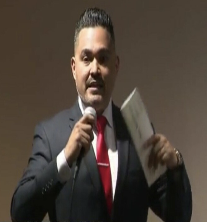 Picture of José Sarmiento in a business suit with red tie and white collared shirt with microphone in hand and papers in the other against a warm grey brownish background