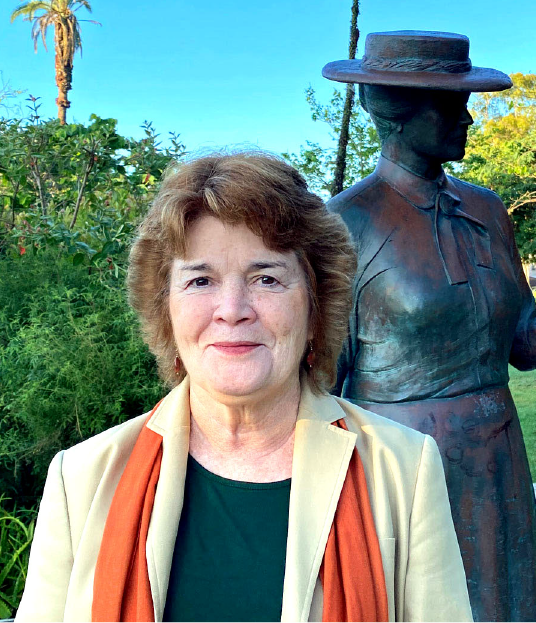 Picture of Kate Callen in a garden with a bronze statue of an early 20th century woman in a wide brimmed hat in the background