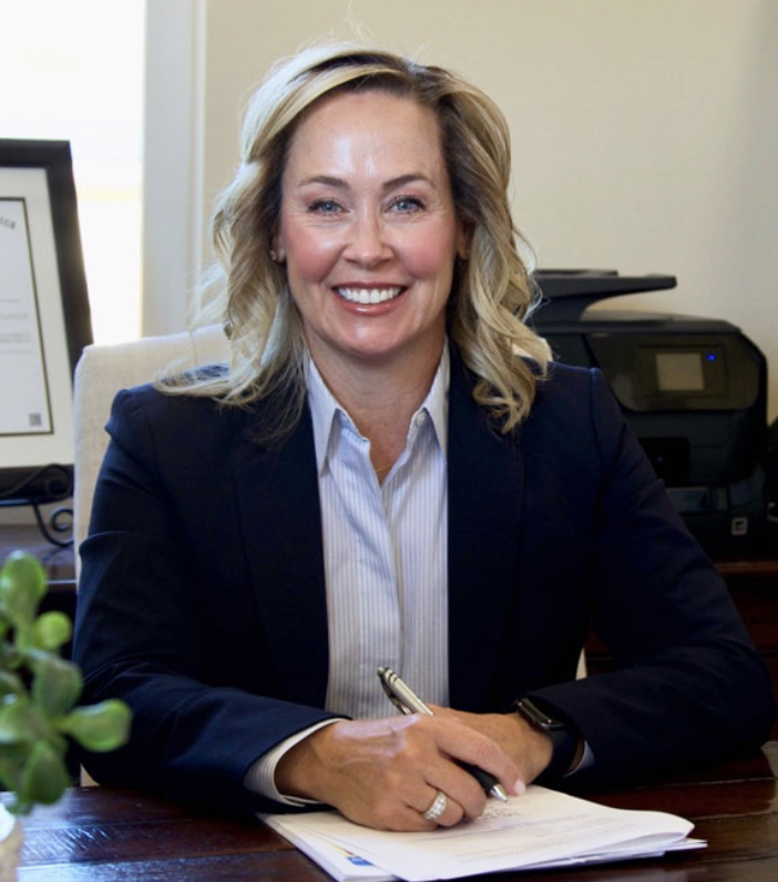 Picture of Kristie Bruce-Lane smiling at a desk with pen in hand wearing a dark navy blue suit jacket and light blue pin-striped collared shirt
