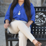 Full body shot of Lauren Cazares sitting crossed-legged and smiling on a La Mesa public bench with blue top and beige pants