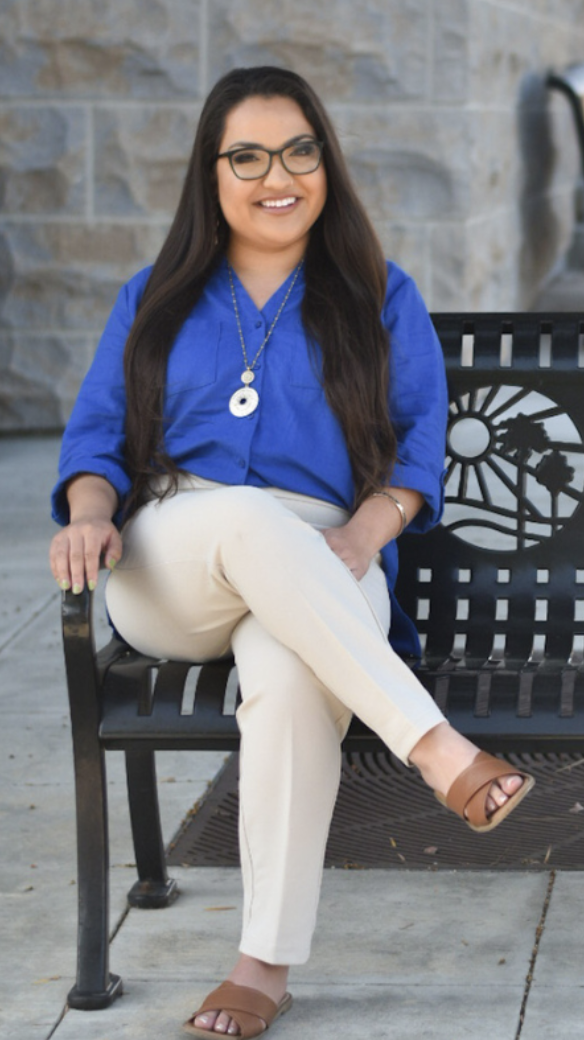 Full body shot of Lauren Cazares sitting crossed-legged and smiling on a La Mesa public bench with blue top and beige pants