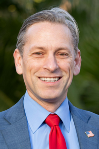 Mark Meuser smiling in a blue business suit with red tie against a blurred greenery background