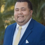 Michael Flemming smiling in a blue business jacket, yellow tie, and white collared shirt against a palm tree background