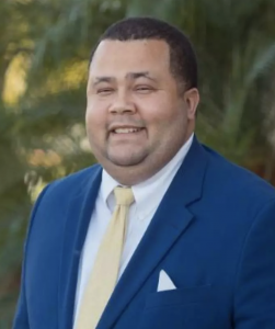 Michael Flemming smiling in a blue business jacket, yellow tie, and white collared shirt against a palm tree background