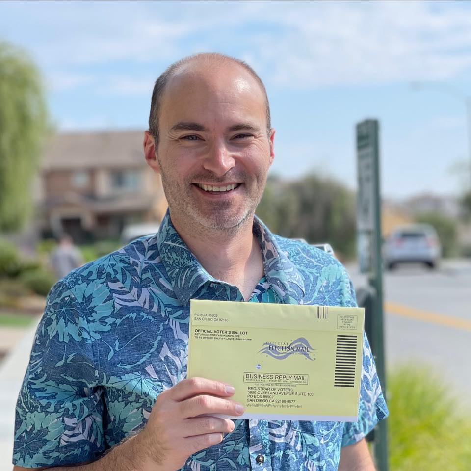 Picture of Paul Mendoza smiling on a bright sunny day in a blue and cyan Hawaiian shirt with a yellow voting ballot envelope in their hands