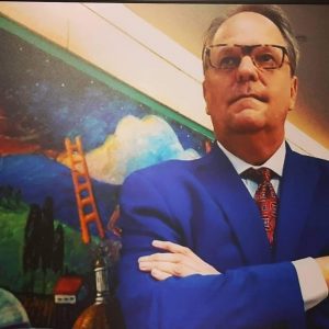 Randy Economy with folded arms in a blue business suit and red tie with a mural in the background