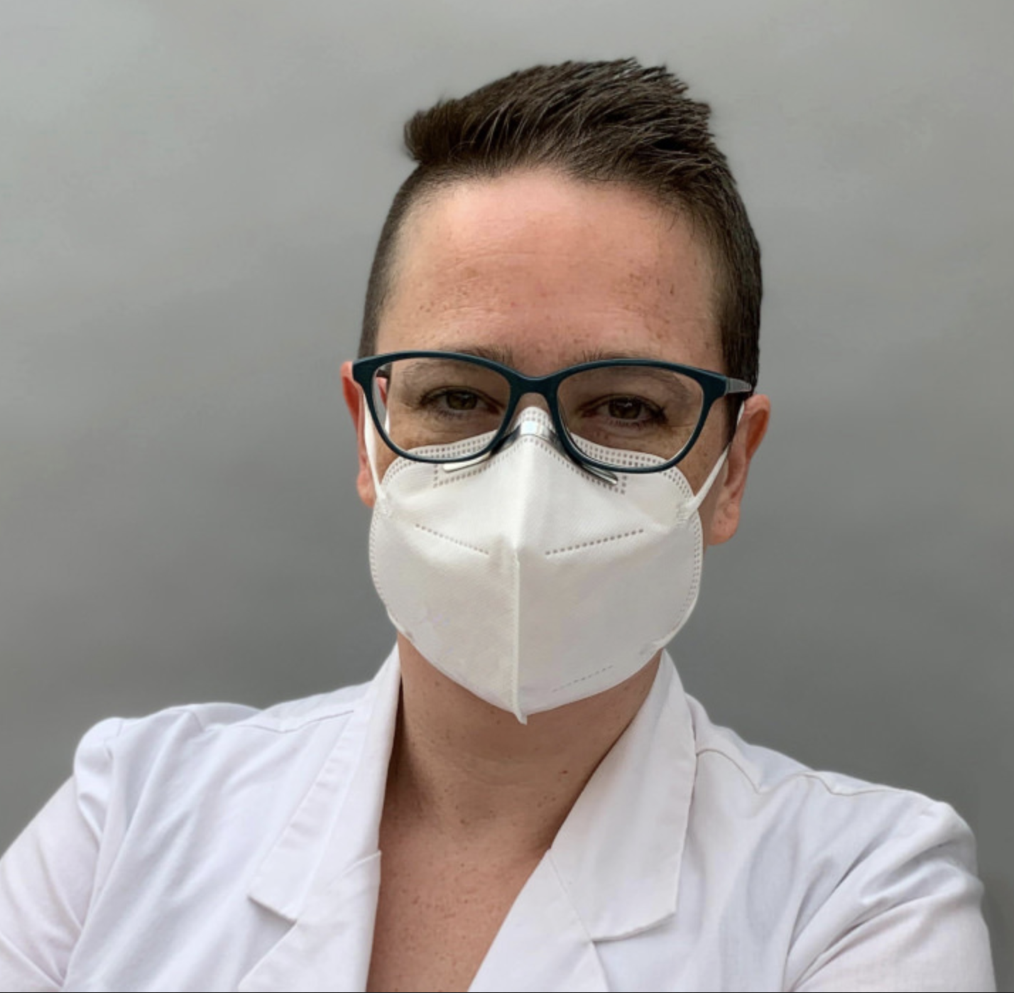 Picture of Sarah Davis in glasses and smiling beneth a white face mask and wearing a medical white coat against a light grey wall